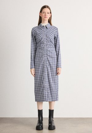 Långärmad, rutigt klänning i blått och vitt med knappstängning, samlad midja och med en strukturerad krage. Svarta ankelstövlar accentuerar looken.