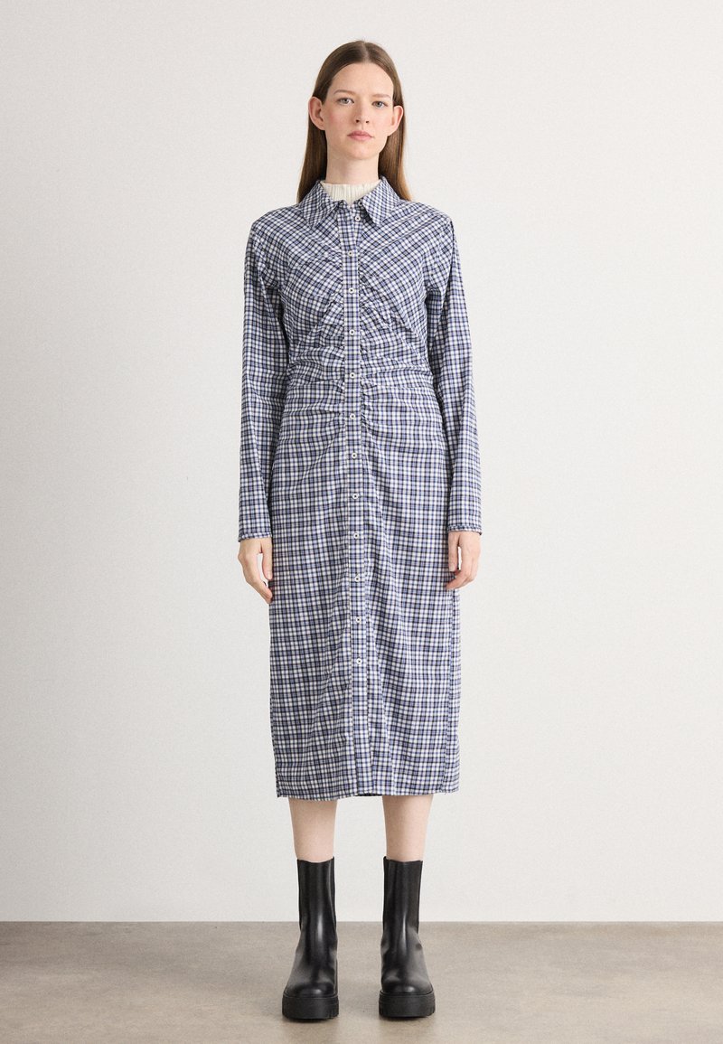 Abito a maniche lunghe, a quadri blu e bianchi con chiusura a bottoni, vita arricciata e colletto strutturato. Stivali neri alla caviglia completano il look.