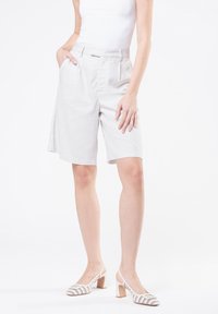 Pantaloni corti grigi chiari su misura con finitura testurizzata, tasche laterali e vestibilità rilassata, abbinati a décolleté bianche con tacco a blocco.