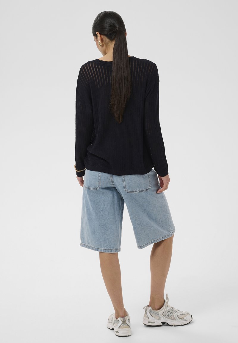 Femme aux longs cheveux foncés attachés en queue de cheval, portant un pull en laine noir, un short en jean bleu clair aux genoux et des baskets blanches, debout de dos.