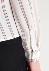 Blusa a strisce con base bianca, linee verticali rosa e nere, e polsini bottonati. Il tessuto appare leggero con una morbida texture.