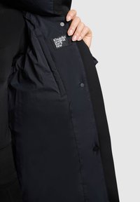 Schwarze isolierte Jacke mit einem glatten, glänzenden Innenfutter und einem Stoffetikett. Verfügt über Druckknöpfe und eine kontrastierende, strukturierte Außenschicht.
