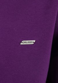 Tela morada con una textura suave y un patrón acanalado, que presenta una etiqueta metálica estampada con "SMILODOX".