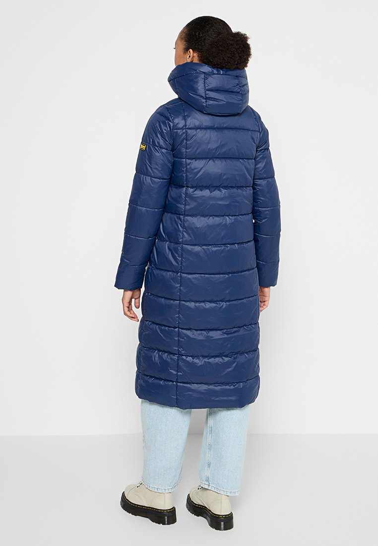 Lungo cappotto voluminous blu navy con colletto alto e cappuccio. Presenta sezioni trapuntate orizzontali e una toppa con logo sulla manica sinistra.