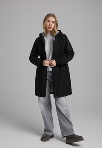 Zwarte lange jas met een capuchon, met ritszakken en elastische boorden, gedragen over een grijze sweater en lichtgrijze spijkerbroek. Gestructureerde stof.