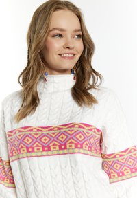 Valkoinen kaapeli-neulepusero, jossa on vaakasuora kirkkaan pinkki ja keltainen geometrinen kuvio rinnassa ja hihoissa. Korkea kaulus.