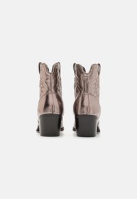 Un par de botas de vaquero metálicas color oro rosa con tacones cuadrados negros, costuras decorativas y lengüetas, vistas desde atrás sobre un fondo blanco.