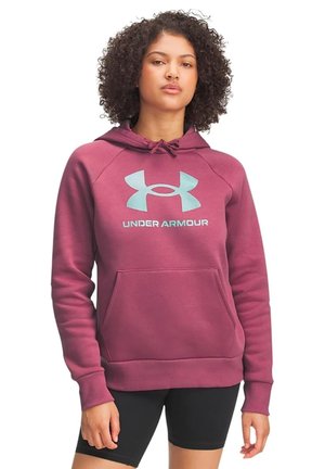 Modèle portant un sweat à capuche rose poudré Under Armour avec une poche avant et un short de cyclisme noir, sur fond blanc.