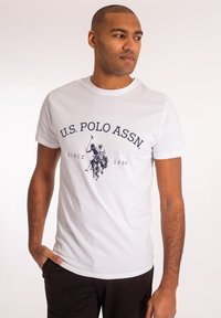 Baltas medvilninis marškinėliai su tamsiai mėlynu spausdiniu, pavaizduojančiu du polo žaidėjus, prekės ženklas "U.S. Polo Assn." ir "Nuo 1890 metų", apvalus kaklas, trumpi rankoviai.