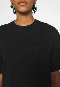 T-shirt noir en tissu léger avec un col rond et des manches courtes. Dispose d'une petite poche sur la poitrine du côté gauche.