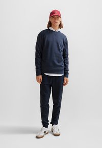 Navy blue sweatshirt with v-neck, matching sweatpants, and white shirt underneath. Red cap with logo and white sneakers with beige accents.
