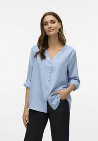 Svetlo modra srajca s kratkimi rokavi in V-izrezom, z zavihanimi rokavi, ki ima teksturirano blago in diskretne gumbe po sprednji strani.