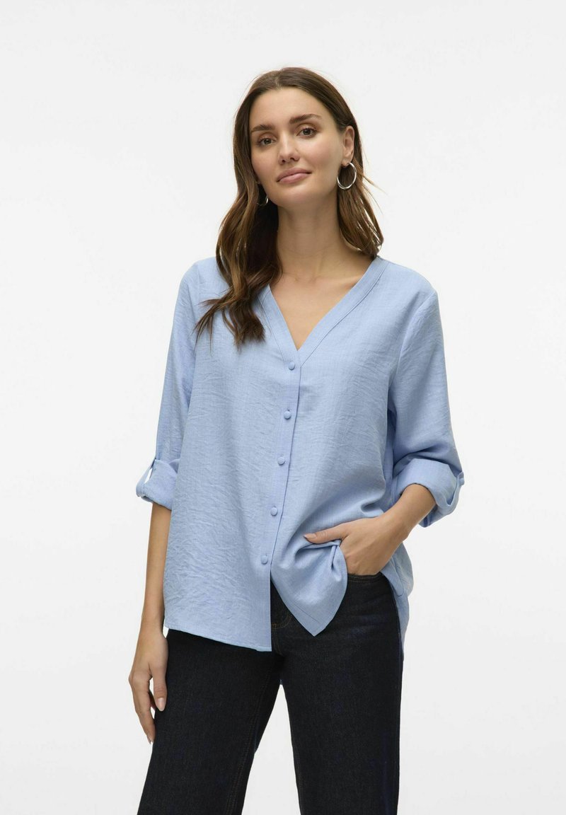 Svetlo modra srajca s kratkimi rokavi in V-izrezom, z zavihanimi rokavi, ki ima teksturirano blago in diskretne gumbe po sprednji strani.
