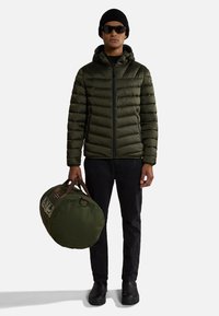 Uomo che indossa un berretto nero, occhiali da sole, giacca a vento verde, pantaloni neri, mentre tiene una borsa da viaggio verde con manici marroni su uno sfondo bianco.