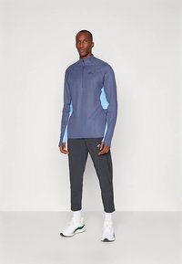 Mężczyzna stoi, ubrany w niebieską sportową bluzę z długim rękawem z jasnoniebieskimi bocznymi panelami, czarne zwężane spodnie, białe skarpetki i białe trampki.