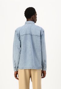 Ανοιχτό μπλε τζιν πουκάμισο με μακριά μανίκια, γιακά, κουμπιά στους καρπούς και ραφή στη πλάτη. Απαλή υφή, χαλαρή εφαρμογή.