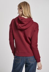 Felpa rossa con cappuccio a cono e vestibilità rilassata, caratterizzata da polsini a coste e cappuccio con cordino. Il materiale sembra morbido con una superficie liscia.