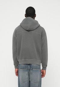 Grauer Kapuzenpullover mit einem kurzen Design, lockerer Passform und Kontrastnähten. Kombiniert mit blauen Jeans in lässiger Passform mit großen Hintertaschen.