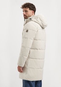 Man in een beige gewatteerde jas met capuchon staat zijwaarts, kijkt naar links, draagt een spijkerbroek tegen een effen witte achtergrond.