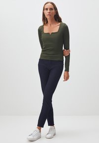 Olivengrünes Langarmoberteil mit quadratischem Ausschnitt, kombiniert mit dunkelblauen, figurbetonten Jeans und weißen Sneakern, vor einem hellen Hintergrund stehend.
