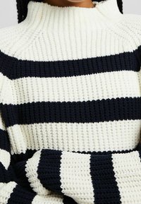 Pletený svetr v krémových a námořnických pruzích. Disponuje vysokým límcem a strukturovanou tkaninou s žebrovanými manžetami a spodním lemem.