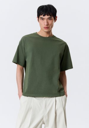 Ung mand stående med hænderne i lommerne iført olivengrøn oversized t-shirt og løse hvide bukser mod en ensfarvet baggrund.