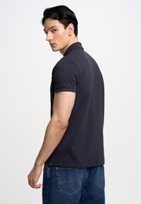 Jūrinės mėlynos spalvos polo marškiniai su trumpomis rankovėmis ir apykakle, derinami su mėlynais džinsais. Medžiaga atrodo glotni ir šiek tiek tekstūruota.