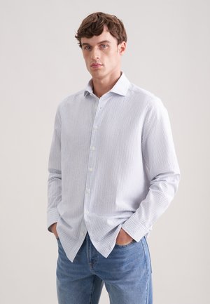 Junger Mann trägt ein weißes langärmliges Hemd mit dünnen vertikalen Streifen und blaue Jeans, steht mit den Händen in den Taschen.