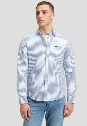 Mann trägt blau-weiß vertikal gestreiftes Langarmhemd über weißem Unterhemd und blauen Jeans, schaut nach links.