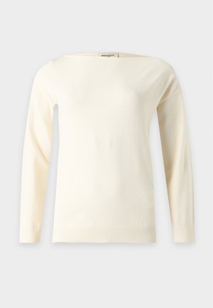 Femme aux longs cheveux foncés debout, vêtue d'un haut crème à manches longues et d'un pantalon large beige avec une ceinture, les mains partiellement dans les poches.