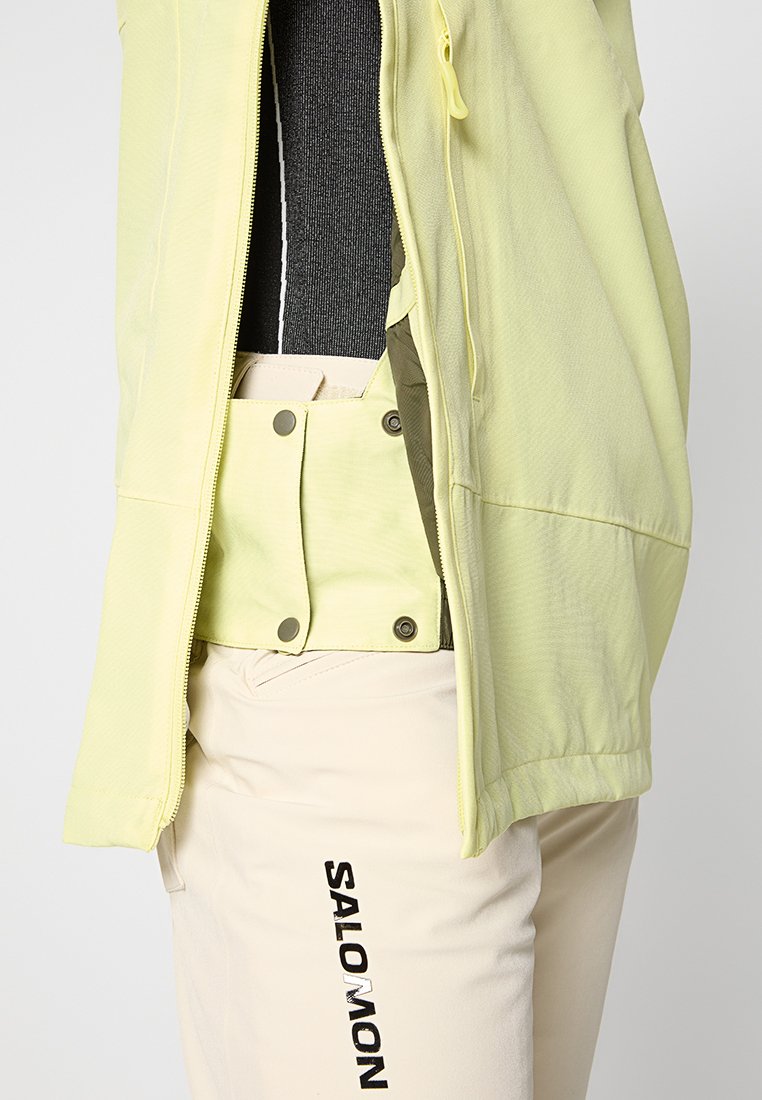 Chaqueta ligera con cremallera en color amarillo pálido, con un acabado texturizado. Combinada con pantalones beige que muestran la marca "SALOMON".