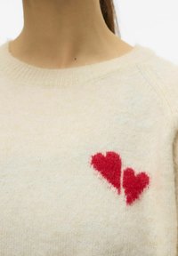 Maglione di lana color crema con scollatura a costine, decorato con due applicazioni a forma di cuore rosso nella zona sinistra del petto, che creano un dettaglio testurizzato.