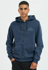 Tamsiai mėlynas zip-up džemperis su raišteliu, priekinėmis kišenėmis ir logotipo spausdinimu. Pagamintas iš minkštos medžiagos, su ribotu rankogaliu ir apačia.