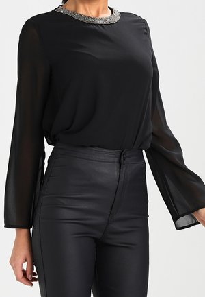 La femme porte un chemisier noir transparent à manches longues avec un col décoratif, associé à un pantalon en cuir noir taille haute.