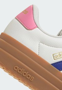 Biały sneaker z teksturowaną skórzaną cholewką, różowymi i niebieskimi akcentami oraz gumową podeszwą w kolorze karmelowym. Na boku znajduje się złote logo "adidas".