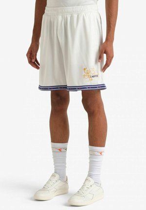 Uomo che indossa pantaloncini sportivi bianchi con rifiniture blu e grafica gialla, calzini bianchi fino al polpaccio e scarpe da ginnastica bianche, in piedi su sfondo bianco.