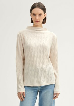Lichtbeige langslijvige top met een opstaande kraag, gekenmerkt door een getextureerd diagonaal ribpatroon. Zacht materiaal met een ontspannen pasvorm.