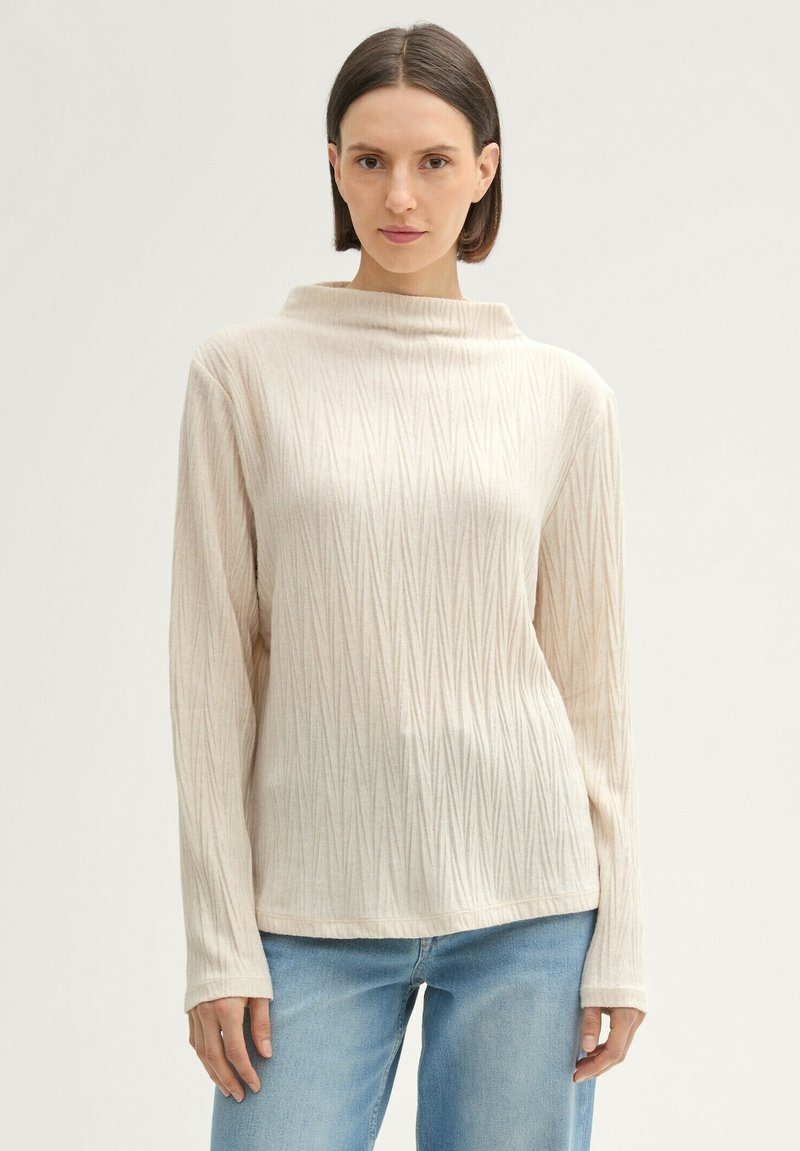 Haut à manches longues beige clair avec un col montant, présentant un motif de côtes diagonal texturé. Matériau doux avec une coupe décontractée.