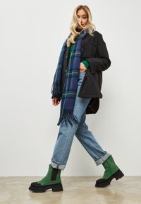 Groene suède enkellaarzen met zwarte accenten, een dikke plateauzool, een blauw geruit sjaal, een zwarte jas en ontspannen blauwe spijkerbroek.