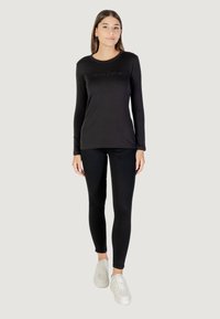 Schwarzes Langarmshirt mit Rundhalsausschnitt und besticktem Logo, kombiniert mit schwarzen Leggings und weißen Turnschuhen. Glatte Stoffstruktur.