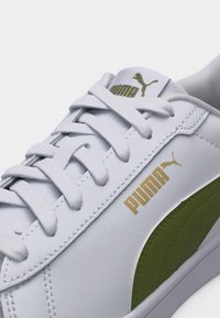Λευκό αθλητικό παπούτσι Puma με πράσινη πλευρική ρίγα, χρυσό λογότυπο Puma στο πλάι και στη γλώσσα, και λευκά κορδόνια δεμένα σε κλασικό σταυρωτό μοτίβο.