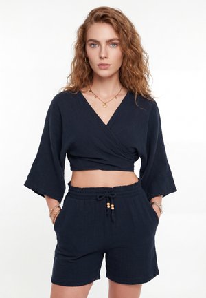 Mladá žena s vlnitými ryšavými vlasmi, ktorá nosí čierny zavinovací crop top a zodpovedajúce šortky so sťahovacou šnúrkou, doplnené zlatými šperkami a hodinkami.