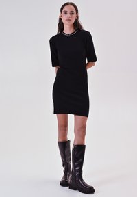 Un vestito nero a maniche corte con scollo decorato, abbinato a stivali neri in pelle al ginocchio con zip laterale e suola a piattaforma.