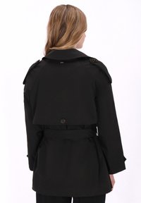 Fekete ballonkabát övvel a derekán, kétsoros gombolású, nagy gallérral és gombos díszítéssel. Simább anyag, elegáns felülettel.