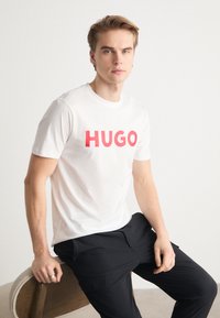 Bílý bavlněný tričko s výrazným červeným textem "HUGO". Model sedí na dřevěné židli a má na sobě černé kalhoty. Jednoduchý design, kulatý výstřih, krátké rukávy.