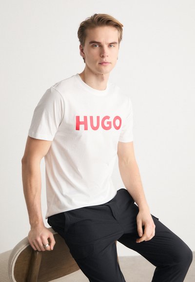 Бяла памучна тениска с голямо червено текстово лого "HUGO". Моделът е седнал на дървен стол, носещ черни панталони. Прост дизайн, с кръгло деколте и къси ръкави.