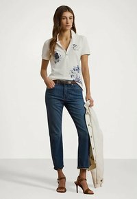 Γυναίκα ντυμένη με λευκό γραφικό πόλο, μπλε τζιν και καφέ ψηλοτάκουνα σανδάλια, κρατώντας μια κρεμ ζακέτα με το ένα χέρι.