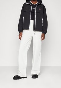 Zwarte ritsjack met capuchon, witte accenten en voorzakken, gedragen over een zwart topje en gecombineerd met witte wijde broek en chunky zwarte laarzen.