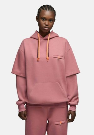 Femme portant un sweat à capuche oversize rose et un pantalon assorti avec des cordons orange et des détails de poche, debout devant un fond uni.