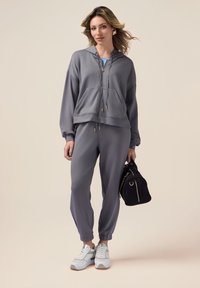 Grauer Hoodie mit Reißverschluss und Kordelzug, kombiniert mit passenden Jogginghosen. Aus weichem Material, mit gerippten Bündchen und lässigem Schnitt. Dazu werden weiße Sneaker getragen.